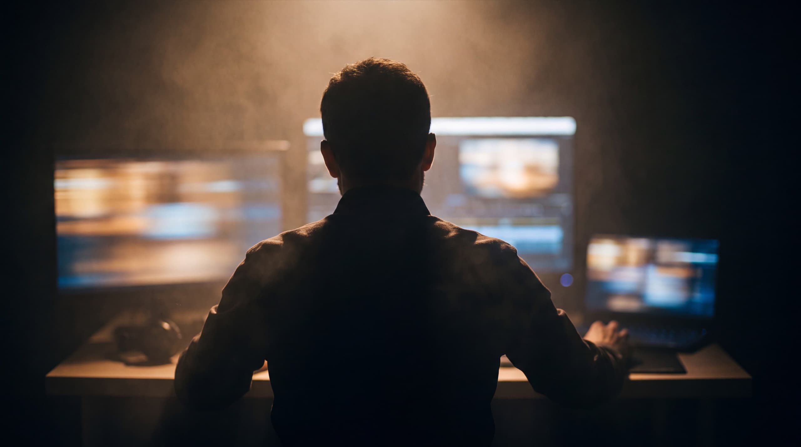 A trader seated at a multi-monitor desk in a darkened room.