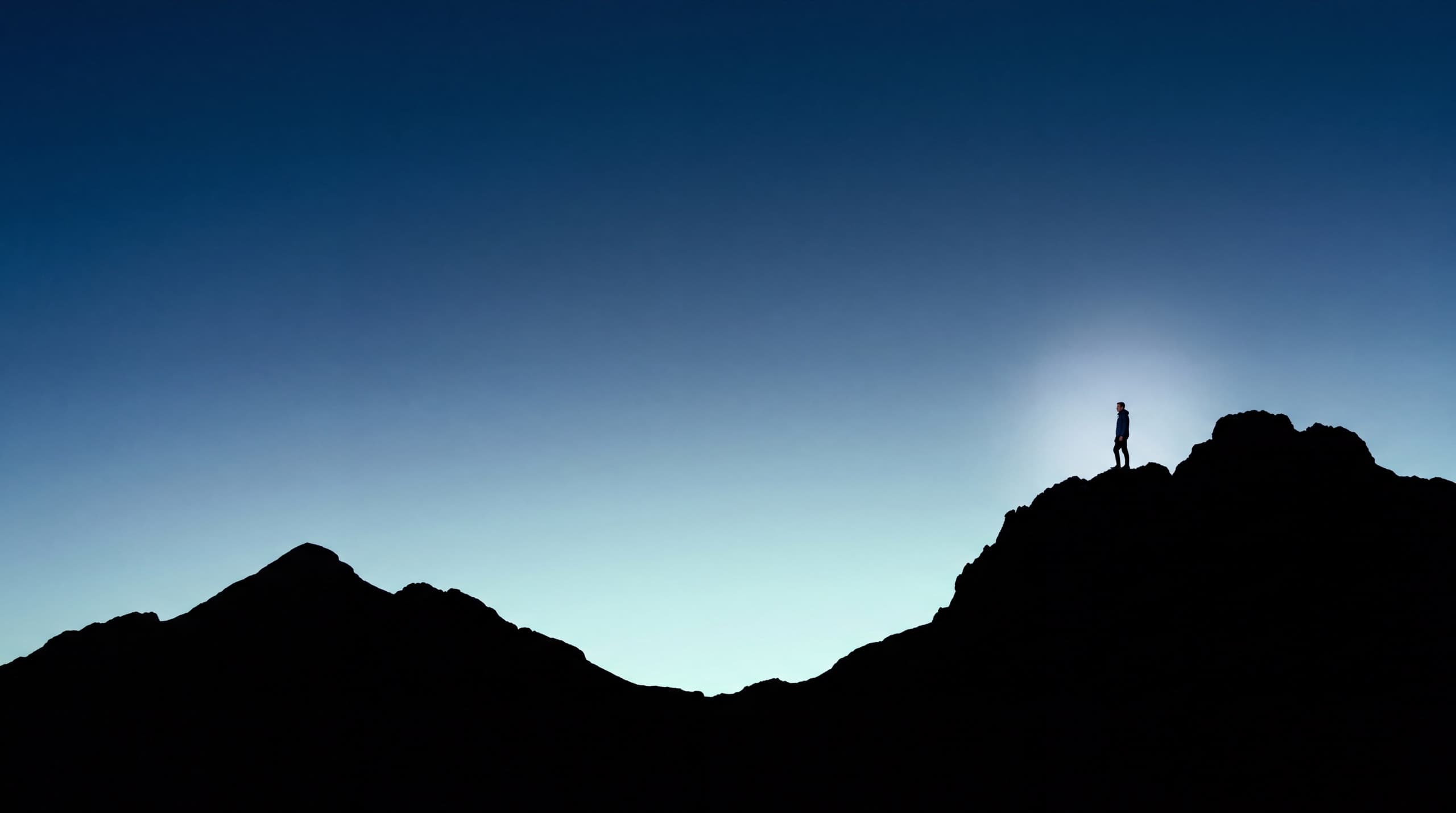 A lone figure standing on the highest summit of a mountain range at blue hour, silhouetted against a pale horizon.